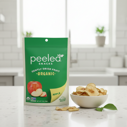 A vibrant green and cream Peeled Snacks Organic Gently Dried Fruit Apple bag, featuring a red apple illustration, stands on a bright white kitchen counter next to a white bowl overflowing with golden-brown dried apple slices and a fresh green apple leaf