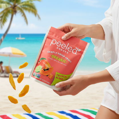 Commercial photograph of hands holding a Peeled Snacks Organic Tropical Blend bag from which dried mango pieces are falling onto a colorful striped beach towel set against a tropical beach with a distant palm tree, turquoise ocean, and a sailboat