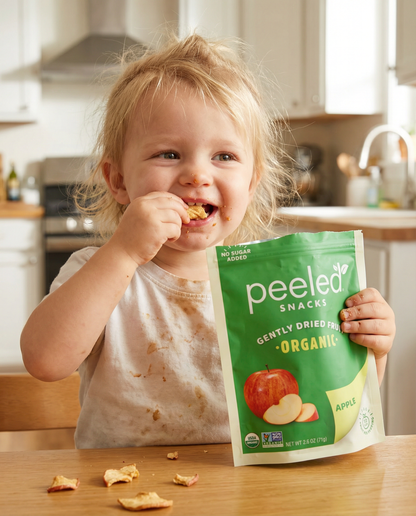 A joyful blonde toddler with a messy face and stained white shirt smiles while eating organic dried apple pieces from a bright green Peeled Snacks bag at a wooden kitchen table