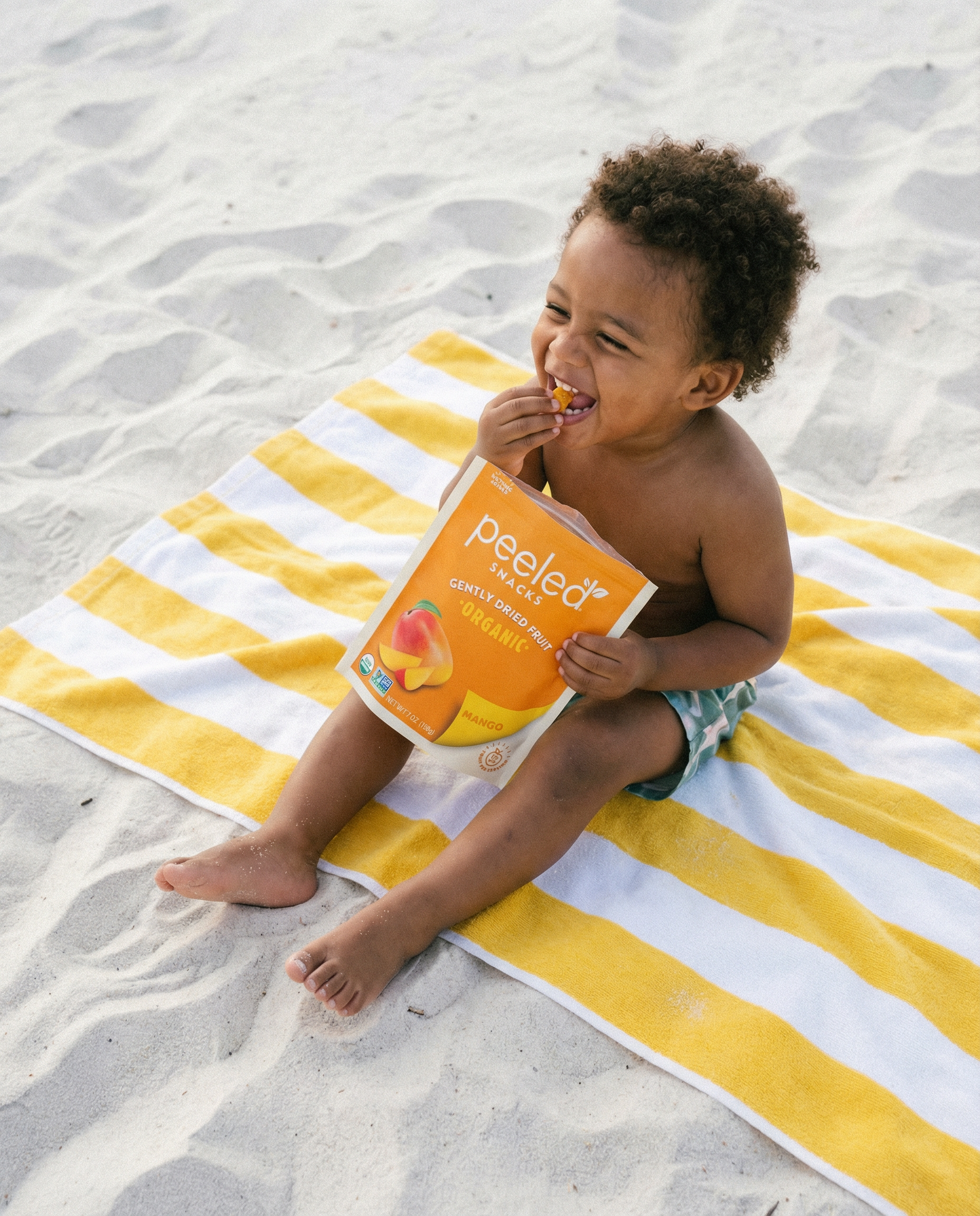A joyful toddler with curly hair sits laughing on a yellow and white striped towel on a white sandy beach, eating a snack from an orange Peeled Snacks Organic Mango pouch