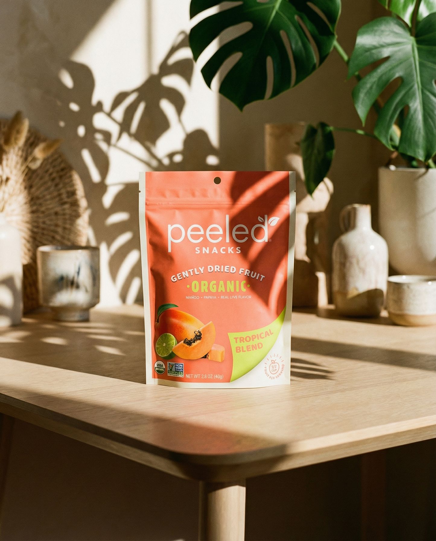 A Peeled Snacks Gently Dried Fruit Organic Tropical Blend salmon-pink pouch with mango, papaya, and lime illustration on a light wooden table, illuminated by dappled sunlight and monstera plant shadows, alongside neutral-toned ceramic vessels