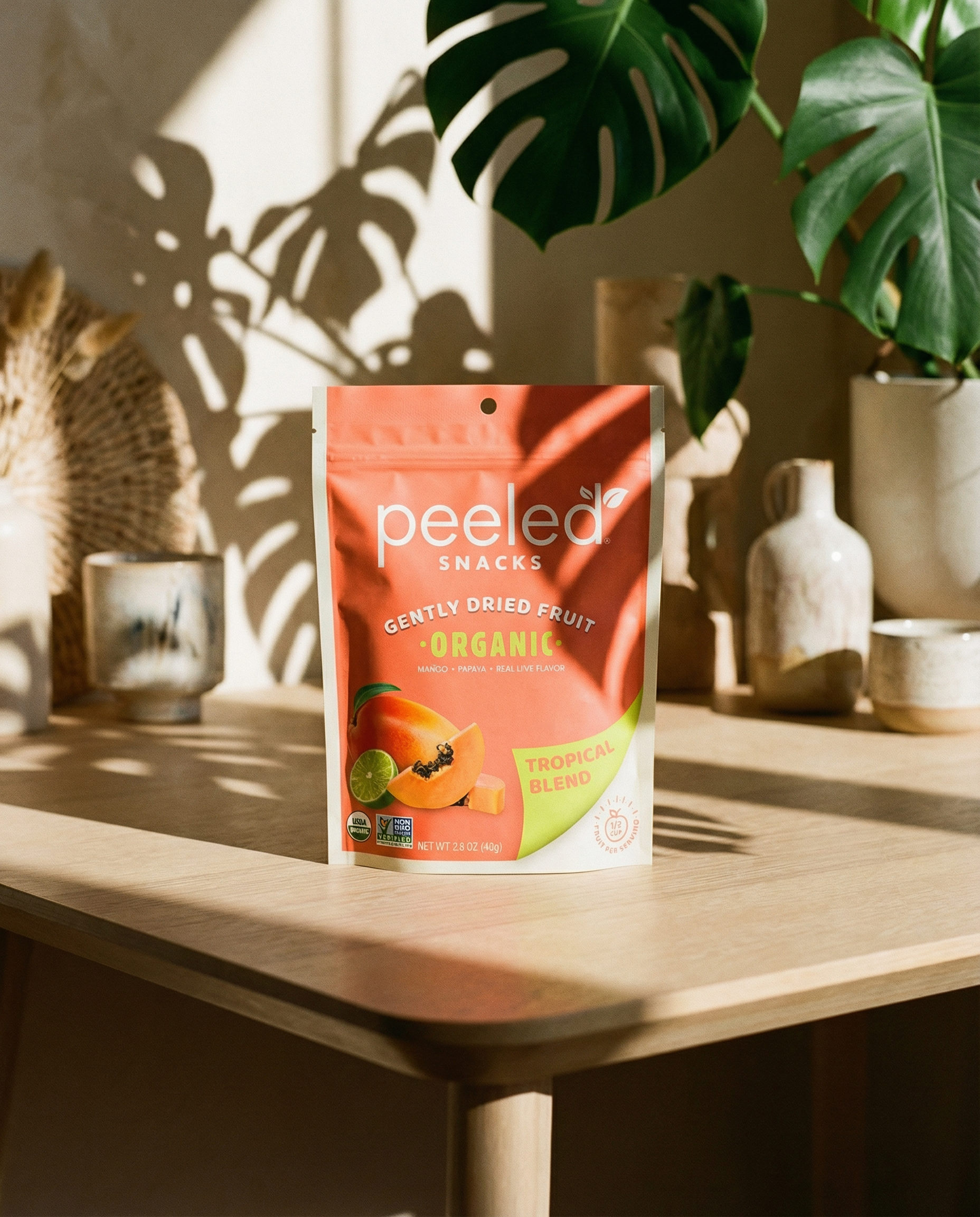 A Peeled Snacks Gently Dried Fruit Organic Tropical Blend salmon-pink pouch with mango, papaya, and lime illustration on a light wooden table, illuminated by dappled sunlight and monstera plant shadows, alongside neutral-toned ceramic vessels