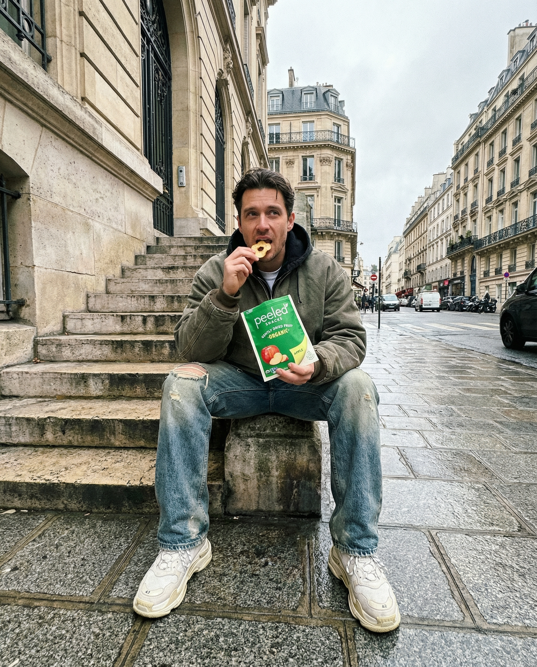 A man in a green jacket, ripped blue jeans, and white Balenciaga sneakers eats a Peeled Snacks Organic Apple chip, sitting on stone steps in a wet Parisian street flanked by classic Haussmann buildings with ornate ironwork and mansard roofs