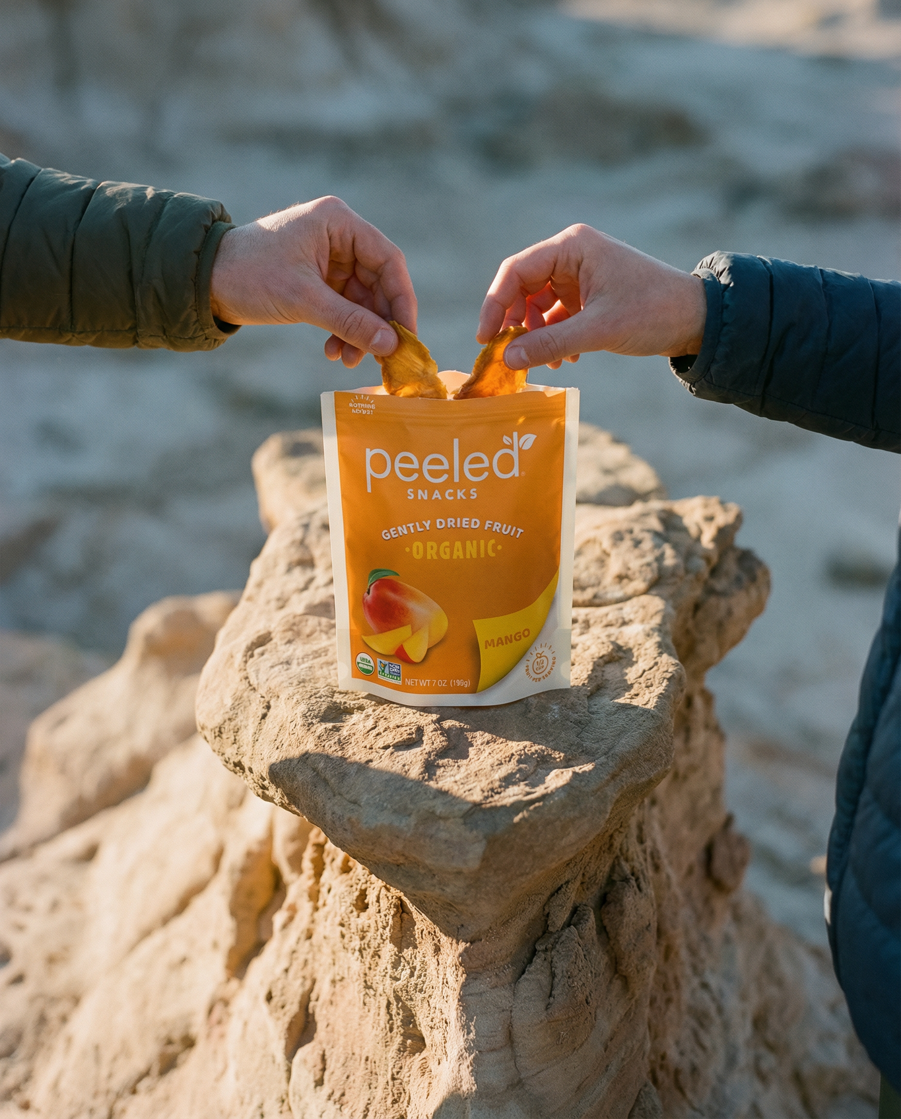 Photograph of two hands, one in a green puffer jacket and one in a blue puffer jacket, sharing golden dried mango slices from an orange Peeled Snacks Organic Mango pouch set on light brown eroded sandstone formations