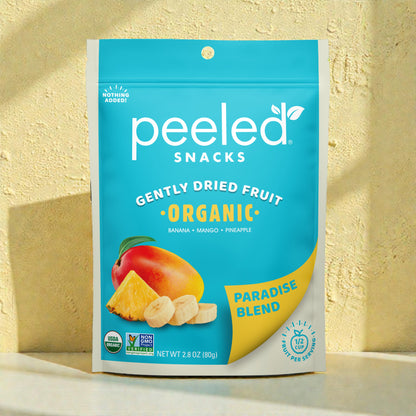 A bright blue Peeled Snacks Gently Dried Fruit Organic Paradise Blend pouch featuring a graphic of mango, pineapple, and banana slices stands on a light beige surface against a textured yellow wall