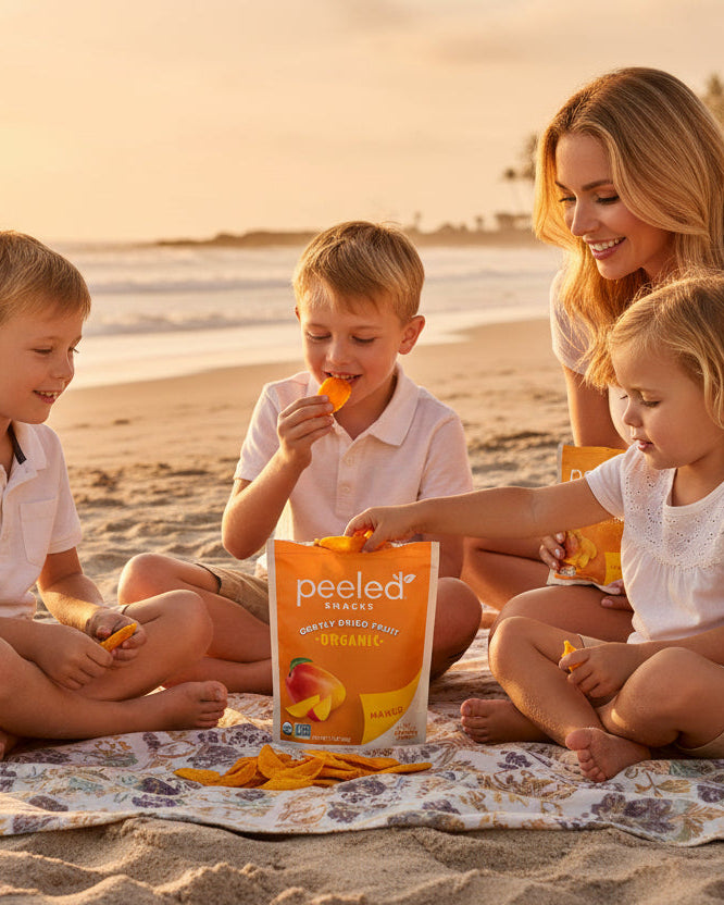 Photography of a mother and three young children happily eating Peeled Snacks organic dried mango on a patterned beach blanket at sunset with palm trees in the distance