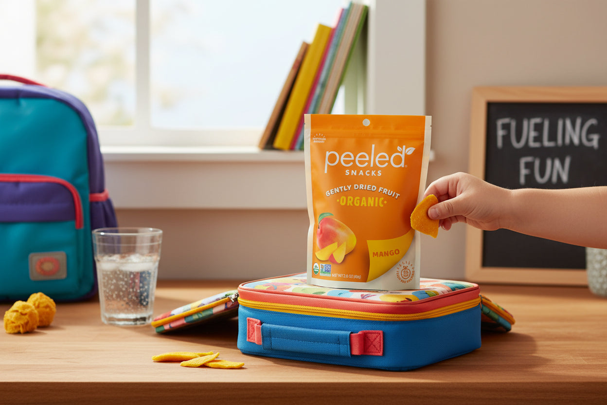 A child's hand holds a piece of dried mango from an orange Peeled Snacks Organic Mango package on a blue and orange patterned lunchbox, with a teal and purple backpack and a chalkboard reading FUELING FUN in a bright room