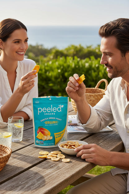A smiling man and woman enjoy Peeled Snacks Organic Paradise Blend dried fruit, holding bright orange mango pieces, at a wooden picnic table with sparkling lemon water and a scenic ocean view