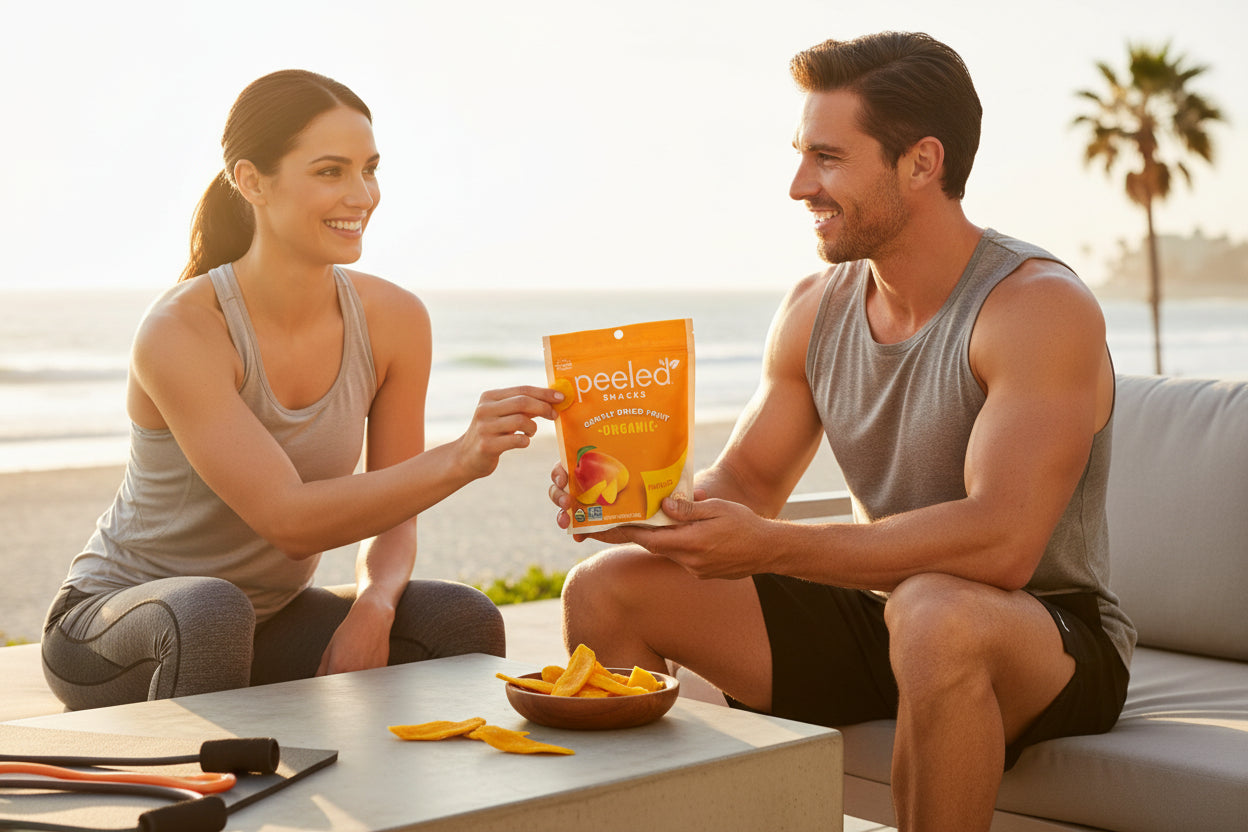 A smiling man and woman in athletic wear share an orange Peeled Snacks organic dried mango pouch and a bowl of yellow mango slices on an outdoor table overlooking a sunny beach with a palm tree in the background and resistance bands nearby