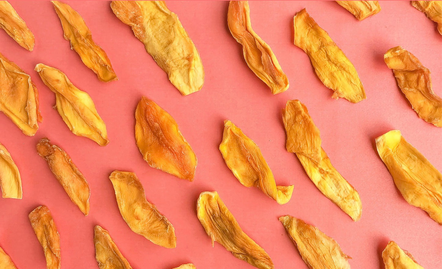 Photograph of golden yellow and orange dried mango slices with varied textures scattered on a vibrant coral pink background