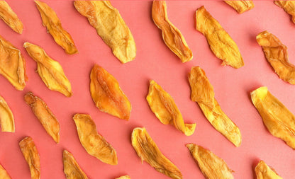 Photograph of golden yellow and orange dried mango slices with varied textures scattered on a vibrant coral pink background